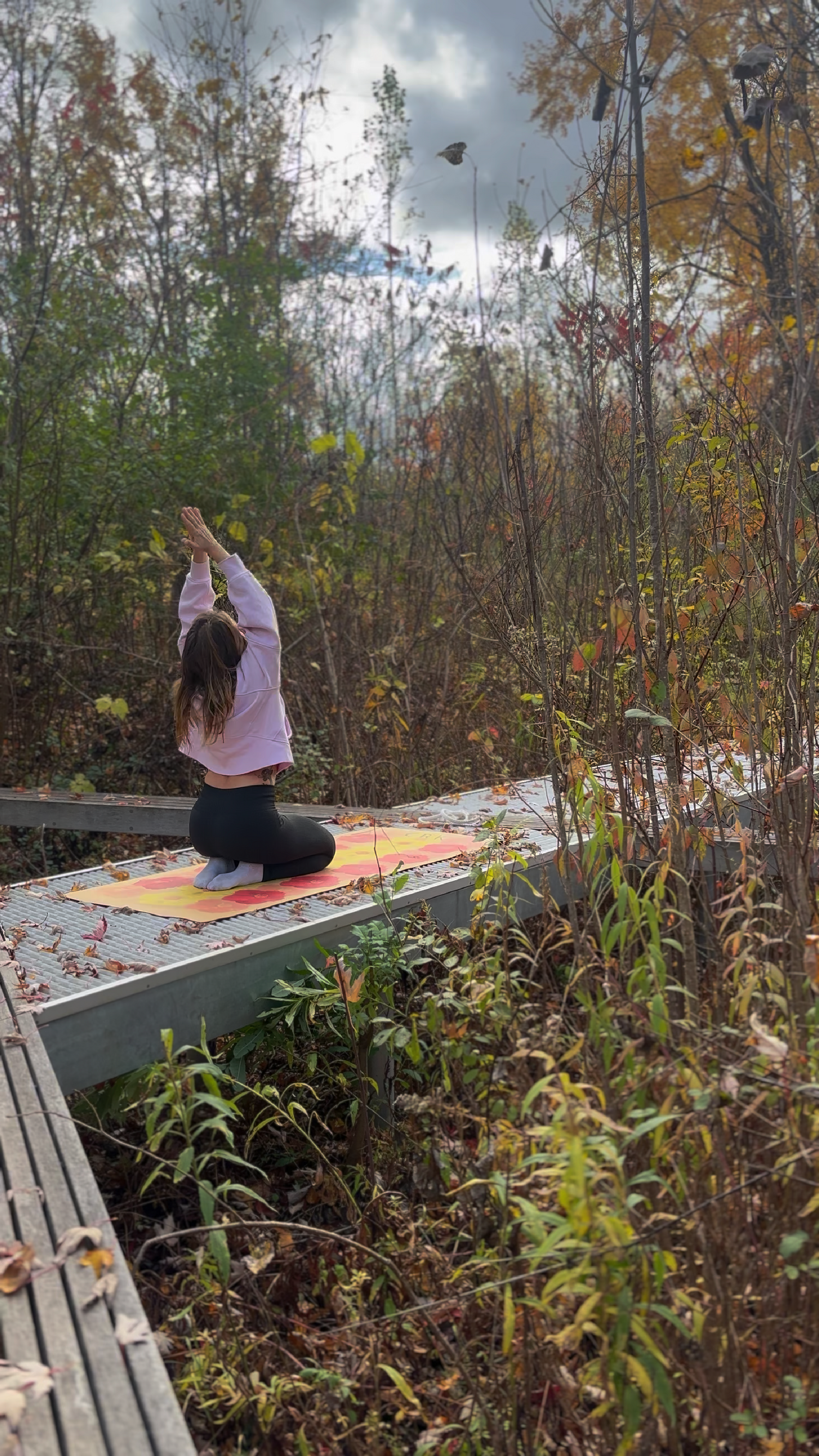 girl in a pink sweater on a path inside  forest on a crk yoga mat with hands in prayer above her head, seated