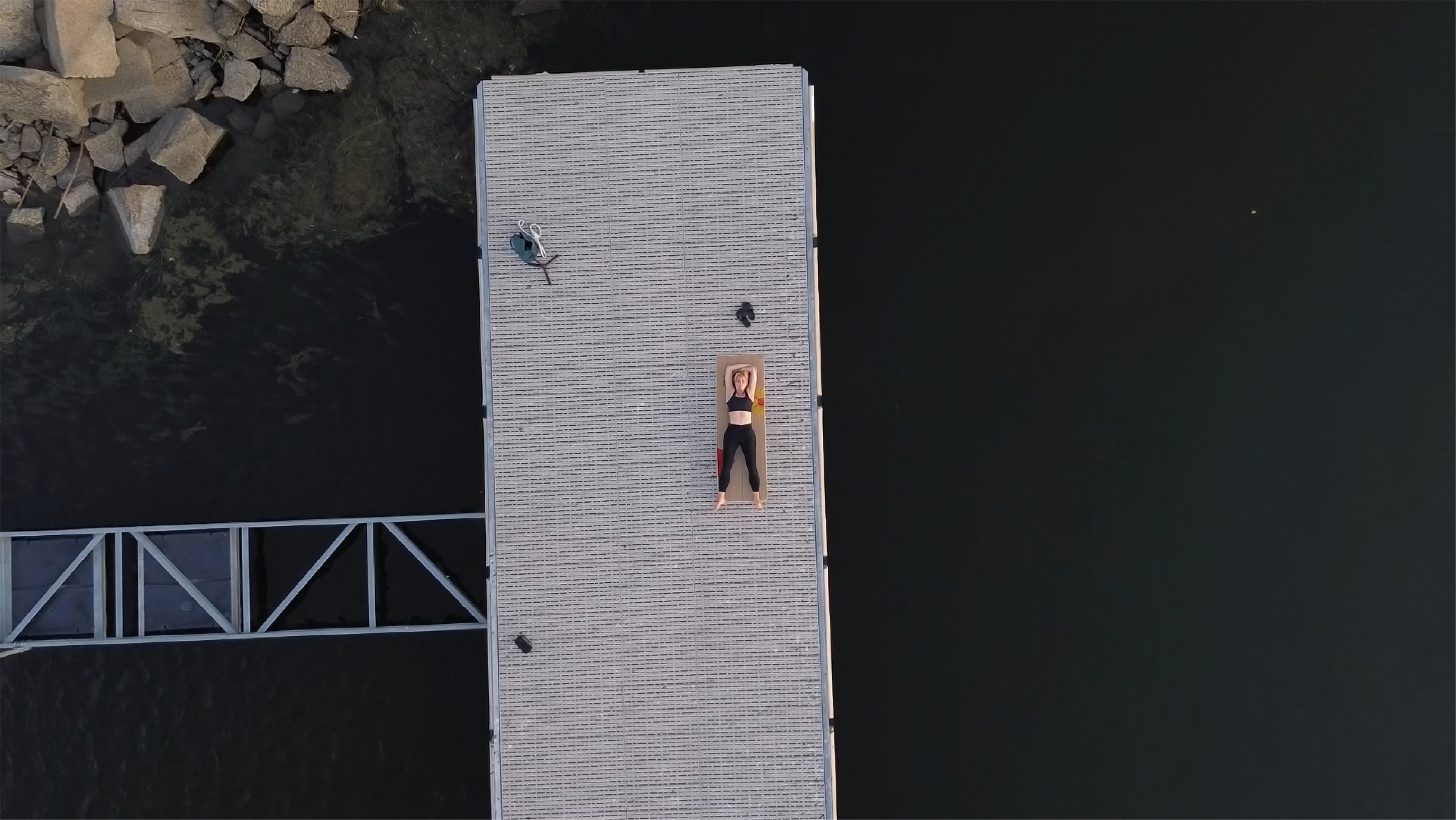 Drone shot at a distance of a girl lying in savasana on her crk yoga mat called the surf mat, made for outdoor yoga practice. she is on a massive metal dock surrounded by nothing but water